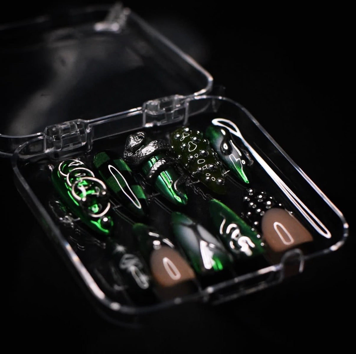 A set of green and black stiletto-shaped press-on nails with silver, black, green and white graphic designs, displayed inside a clear plastic case on a black background.