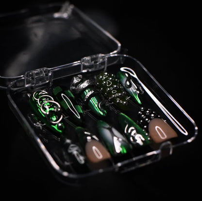 A set of green and black stiletto-shaped press-on nails with silver, black, green and white graphic designs, displayed inside a clear plastic case on a black background.
