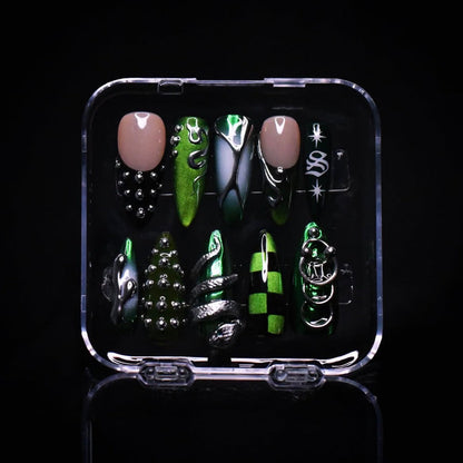 A set of coffin-shaped press-on nails displayed in a clear plastic case against a black background. The nails feature a mix of metallic green, black, and silver designs with gothic and fantasy motifs.
