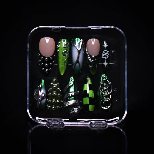 A set of coffin-shaped press-on nails displayed in a clear plastic case against a black background. The nails feature a mix of metallic green, black, and silver designs with gothic and fantasy motifs.