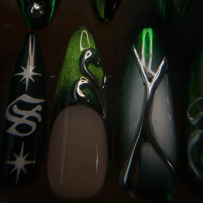 Close-up of several artificial nail tips displayed on a dark surface, featuring glossy black, metallic green, and silver swirl details, and white starburst graphics.