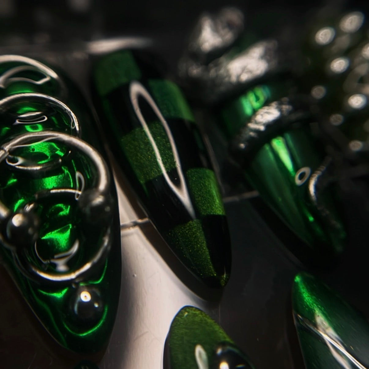 A close-up of glossy green and black checkered false nails with metallic silver rings, displayed on a reflective surface.