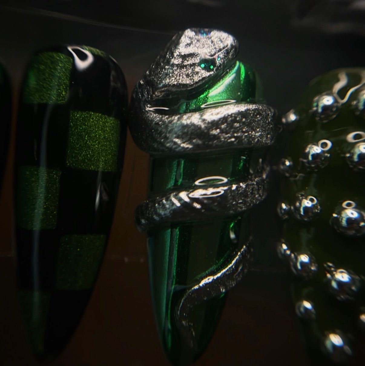 A close-up of decorative nail art featuring a metallic silver snake wrapped around a glossy emerald green nail tip, alongside other green and black nail designs in the background.